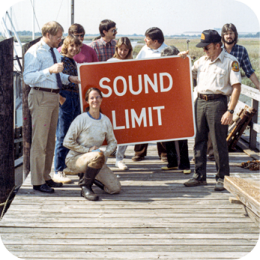 CRD manages, protects, and enhances the state's coastal and marine resources. Through research, policy, and public engagement, CRD supports sustainable use and conservation of Georgia's coastal ecosystems. Learn more about our division here. A group of people, including men and women dressed in casual and uniformed attire, stand on a wooden dock holding a large red sign that reads "SOUND LIMIT." One woman, wearing work clothes and boots, kneels in front of the sign while smiling at the camera. The background features a coastal marsh with tall grasses and calm water under a clear sky. Some individuals in the group are engaged in conversation, while others look at the sign or the camera.