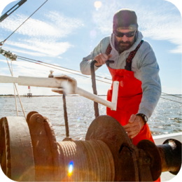 Coastal Resources Division provides regulations, licensing information, and resources for commercial fishing along the coast. It covers permitted species, fishing regulations, and sustainability practices to support the fishing industry.  A commercial fisherman wearing a gray hoodie, red waterproof overalls, sunglasses, and a black beanie is working on a fishing boat. He is operating a large metal winch with one hand while gripping a lever with the other. The sun shines brightly, creating a lens flare effect. The background features open water with a distant structure visible on the horizon. The sky is mostly clear with a few wispy clouds.