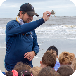 CRD's Education and Outreach Unit engages communities by promoting awareness of Georgia’s coastal ecosystems and conservation efforts. Through interactive programs, field experiences, and public outreach, they inspire stewardship of marine and coastal resources. A male educator wearing a navy blue long-sleeve shirt, black cap, and patterned neck gaiter holds up a small crab while speaking to a group of children on a sandy beach. The children, with their backs to the camera, are attentively watching. The ocean waves roll onto the shore in the background under an overcast sky.