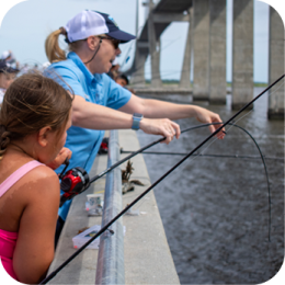 The Coastal Resources Division provides information on fishing regulations, licensing, and conservation efforts for coastal waters. Anglers can find details on fish species, seasonal rules, and sustainable fishing practices. A young girl in a pink swimsuit and an adult in a blue shirt and white cap are fishing from a concrete pier. The adult is pointing and assisting the girl, who is holding a red and black fishing rod. Several other people are fishing in the background, and a large bridge stretches over the water. The water below is calm, and the sky is overcast.