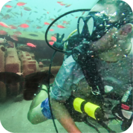 Coastal Resources Division's Habitat Enhancement and Restoration Unit works to improve coastal ecosystems through artificial reef development, oyster reef restoration, and other conservation efforts. These projects support marine life, improve water quality, and enhance fishing opportunities while promoting coastal resilience. A scuba diver wearing a black diving mask, regulator, and a yellow oxygen tank explores an underwater artificial reef. The reef consists of large, hollow concrete structures resting on the ocean floor. Small red fish swim around the diver and the reef. Bubbles rise from the diver’s regulator, and the water has a greenish tint with particles suspended in it.